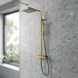 Nuie Square thermostatic fixed head mixer bar shower with brushed brass finish and shower kit installed on modern gray tiled bathroom wall next to window