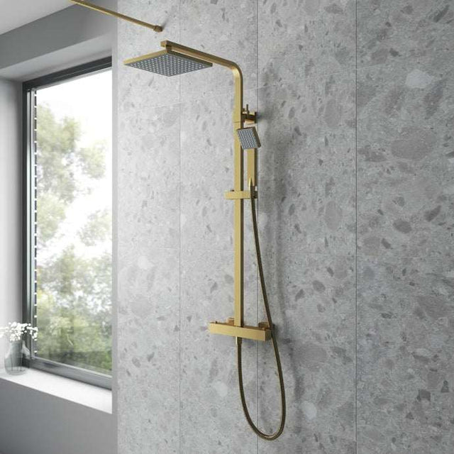 Nuie Square thermostatic fixed head mixer bar shower with brushed brass finish and shower kit installed on modern gray tiled bathroom wall next to window